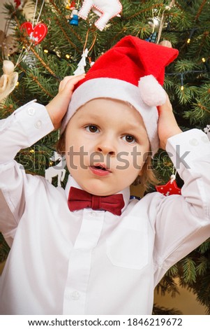 Surprised or shocked baby boy dressed in festive clothing and Santa Claus hat. Christmas tree with decoration in background. Christmas time concept