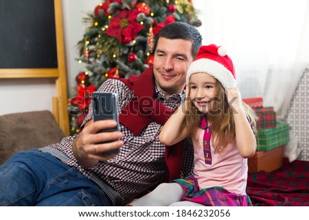 Dad and daughter near the Christmas tree with a smartphone take a selfie, communicate via video connection. Christmas greetings, a gift box, a girl in a Santa hat waving Hello. New year, holiday decor