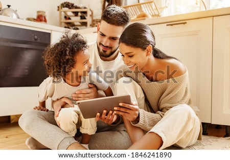 happy multi ethnic family: parents and little son watching cartoon on tablet on weekend day  while sitting on the kitchen floor at home together 

