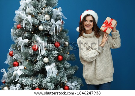 Happy young woman in Santa hat with gift near Christmas tree on blue background