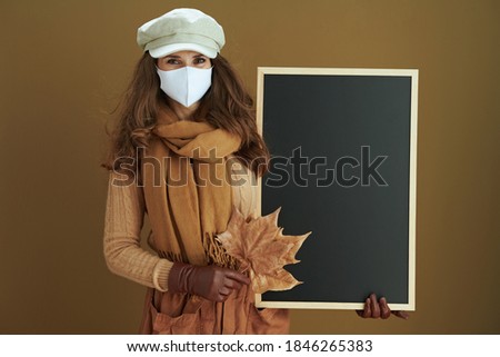 Life during coronavirus pandemic. young female in scarf with white medical mask and yellow autumn maple leaves showing blank board on beige background.