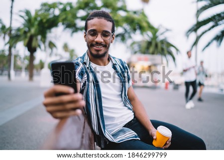 Happy bearded ethnic male in casual outfit and glasses with cup of hot coffee in disposable cup taking selfie while sitting on bench