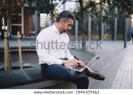 Mature male trader spending time for online business sitting outdoors at urban setting, middle aged entrepreneur using 4g wireless internet connection for uploading web files on modern laptop computer