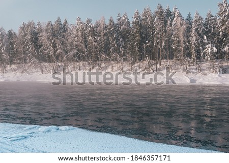 Frosty fog over winter river with snow and forest on bank. First ice on lake on cold day.
