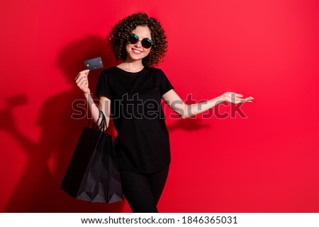 Photo of sweet curly woman wear casual black outfit eyeglasses holding bags credit card pointing empty space isolated red color background