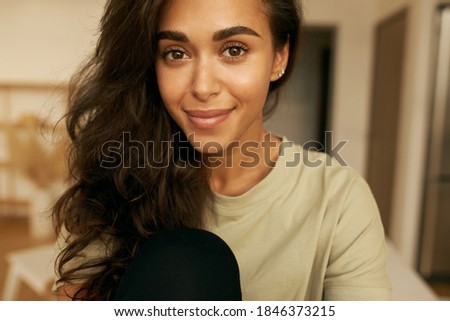 Close up image of attractive young Hispanic female with tanned perfect skin, voluminous hair and brown eyes smiling, being in good mood, spending nice time at home, wearing casual clothes