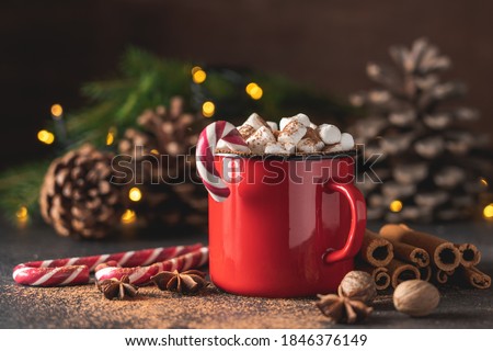 Hot drink with marshmallows and candy cane in red mug. Fir cones, spices in the background. Cozy seasonal holidays