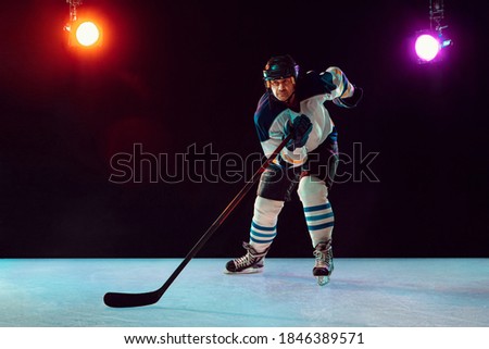 Winner. Male hockey player with the stick on ice court and dark neon colored background. Sportsman wearing equipment, helmet practicing. Concept of sport, healthy lifestyle, motion, wellness, action.