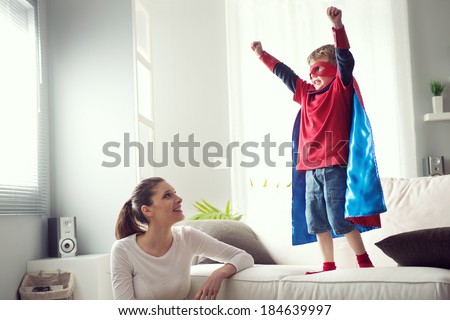 Mother looking at little superhero with fists raised standing on sofa. Royalty-Free Stock Photo #184639997