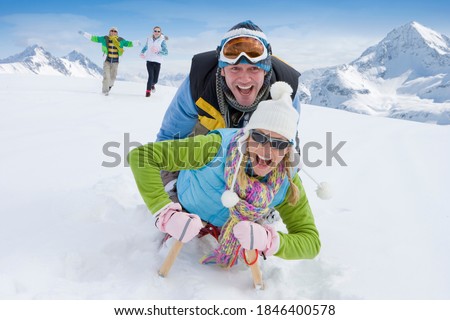Husband and wife together sledding down a steep snowy slope on a single sled with their running in the background to chase them