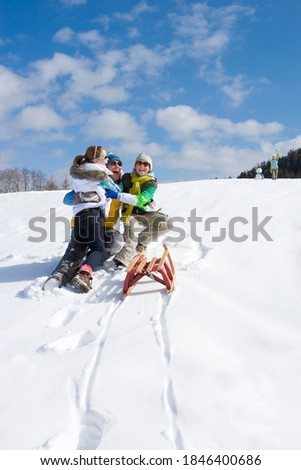 Father happily hugging and holding his kids near the sled on a steep ski slope while mother standing at the top next to a snowman