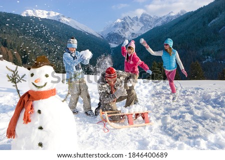 Man sitting down near the sled on a ski slope and being playfully attacked by his family during a snowball fight next to a snowman