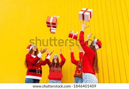 Excited, rejoicing group of friends wearing santa claus hats having fun and tossing christmas presents on yellow wall
