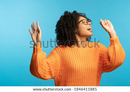 Funny dark skinned millennial woman with curly hair in good mood sings her favorite song, dancing, wear hipster oversize orange sweater, isolated on studio blue background. Mock up, copy space. 