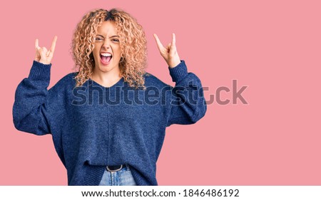 Young blonde woman with curly hair wearing casual winter sweater shouting with crazy expression doing rock symbol with hands up. music star. heavy music concept. 