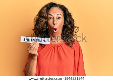 Middle age hispanic woman holding engagement word paper scared and amazed with open mouth for surprise, disbelief face 