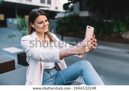Happy millennial woman enjoying leisure time for listening favourite positive music via electronic earphones and mobile application, cheerful hipster girl clicking selfie photo on front camera