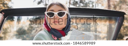 Elegant woman in sunglasses and headscarf holding map in roofless auto, banner