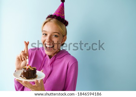 Excited girl posing with birthday cake and holding fingers crossed isolated over blue background