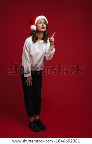 Photo of cute shocked young woman in christmas santa hat isolated over red wall background showing copyspace