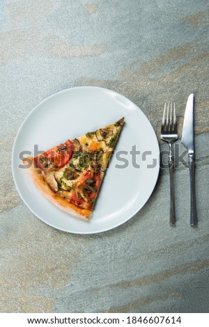 Slice of veggie pizza on plate. Knife and fork near the dish on stone anthracite background. Piece of vegetable pizza with tomatoes, champignon mushrooms, sliced eggplant with pesto sauce. Top view