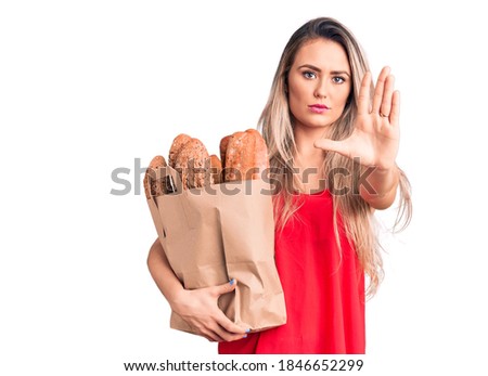 Young beautiful blonde woman holding paper bag with bread with open hand doing stop sign with serious and confident expression, defense gesture 