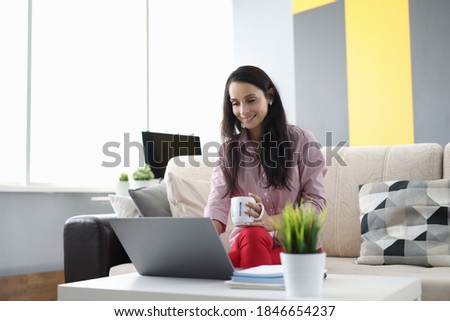 Woman is sitting on couch holding cup in her hands and working at laptop. Remote work at home for freelancers concept