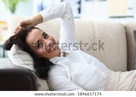 Portrait of happy woman with charming smile looking at camera with happiness. Female person relaxing on comfortable couch. Office interior on background