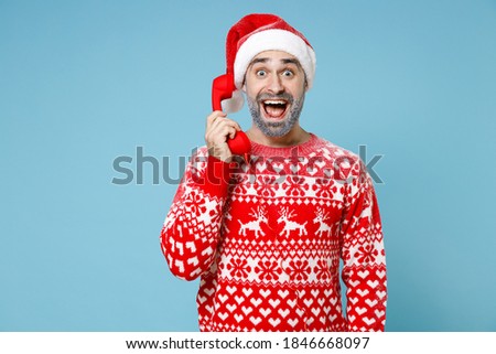 Surprised Northern bearded man frozen icy snow face in Santa hat Christmas sweater talk on telephone isolated on blue background studio. Happy New Year celebration merry holiday winter time concept