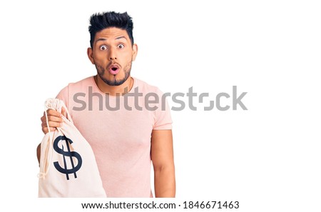 Handsome latin american young man holding money bag with dollar symbol scared and amazed with open mouth for surprise, disbelief face 