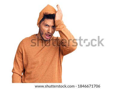 Handsome latin american young man wearing winter sweater and wool hat surprised with hand on head for mistake, remember error. forgot, bad memory concept. 