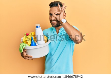 Handsome man with beard holding cleaning products smiling happy doing ok sign with hand on eye looking through fingers 