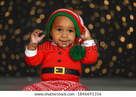 Cute little African American baby wearing elf hat against blurred lights on dark background. Christmas celebration