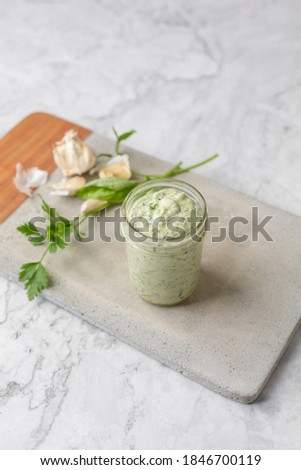 Homemade Green Goddess Dressing in a Jar on a Cement Cutting Board on White Marble Countertop; Parsley and Garlic in Background