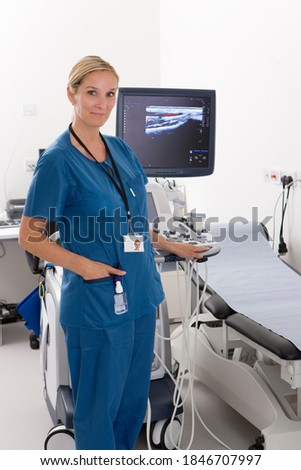 A vertical view of a qualified technician posing for the camera in blue uniform while standing next to the ultrasound machine in a clinic