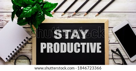 STAY PRODUCTIVE is written in white on a black board next to a phone, notepad, glasses, pencils and a green plant.