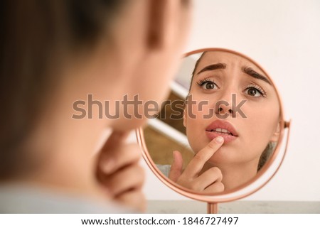 Emotional woman with herpes touching lips in front of mirror against light background Royalty-Free Stock Photo #1846727497