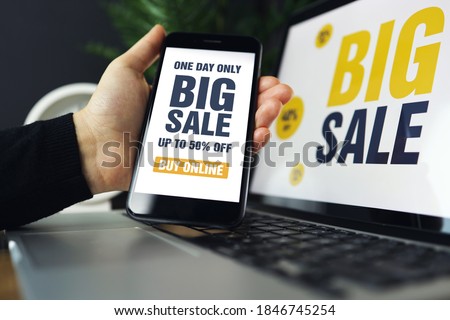 Closeup of smartphone with white screen and sign - Big Sale in female hand. In background blurred notebook. Woman using the gadget to surf Internet, online shopping, store app.