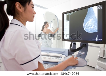 A young adult woman taking the mammogram test on a machine in a clinic while being examined by a professional radiologist in a lab coat through a high definition x-ray monitor Royalty-Free Stock Photo #1846749064