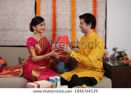 Indian husband surprising wife with a gift on Diwali / anniversary / birthday while sitting on sofa.