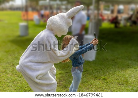 A young girl takes a selfie of the Easter Bunny and her at the community park Easter Festival. The Easter Bunny crouches down behind the child holding up two fingers looking into the smartphone.