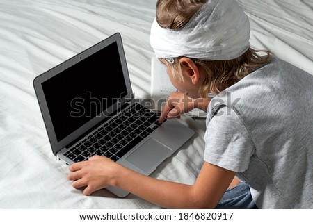 Little Caucasian blond boy with a head injury and bandage is sitting on the bed and using laptop. Recovery after incident. Remote communication, distance learning, technologies and gadgets in life