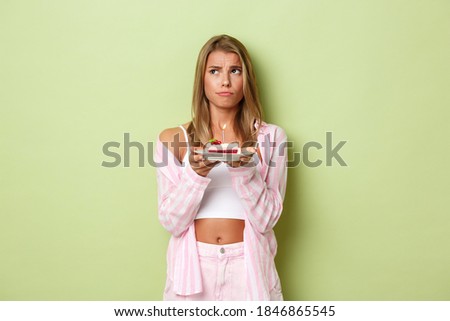 Image of gloomy blond girl with birthday cake, looking at upper left corner, puzzled to make wish, standing over green background