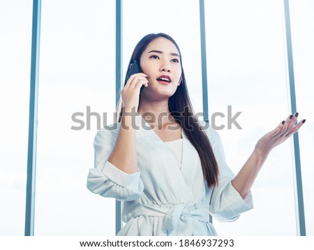 Young pretty Asian woman wearing formal white shirt talking on the phone call near huge glass window in office. Portrait of beautiful business woman using mobile phone in conversation.