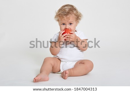 Beautiful blond curly haired little boy eats fresh red apple, healthy breakfast, starts day with healthy food, infant wearing body suit sitting barefoot on floor against white wall.