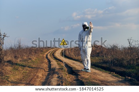 Man in white coverall, gas mask, gloves standing on road in field next to caution symbol showing stop sign, on a sunny day. Biohazard sign warning about harmful biological substances and danger