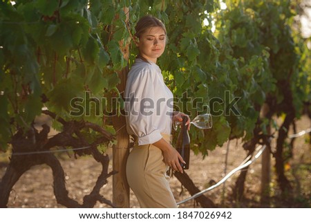 Beautiful woman sommelier checks grapes before harvest. Winner in a white blouse with a bottle of red wine and a glass. Outdoor farmer countryside style