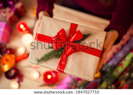 Present box with beautiful red ribbon and small branch of fir-tree in girls hands. Table with christmas decoration on the background, warm atmosphere of the picture.