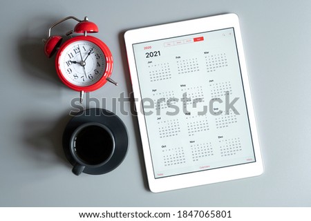 a tablet with an open calendar for 2021 year, a cup of coffee,  and a red alarm clock on a gray background Royalty-Free Stock Photo #1847065801