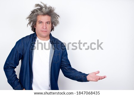 Portrait of Handsome middle aged Caucasian man with afro gray hair standing against white wall  with arm out in a welcoming gesture.